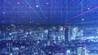 Futuristic cityscape at night with illuminated buildings overlaid by a digital grid of blue and pink lines. Represents smart city technology, data networks, IoT