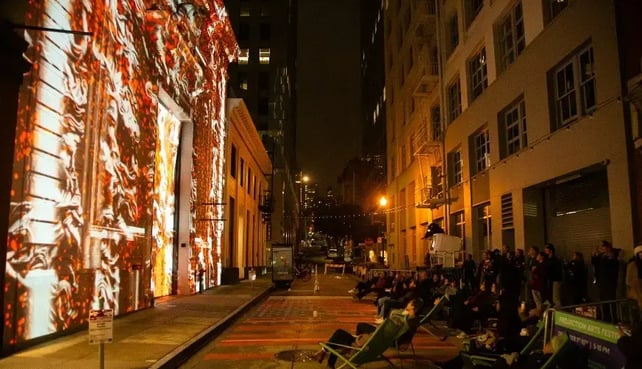 Projection mapping lights up the side of a city building with orange colors as a group of people stand across the street to watch it at night.