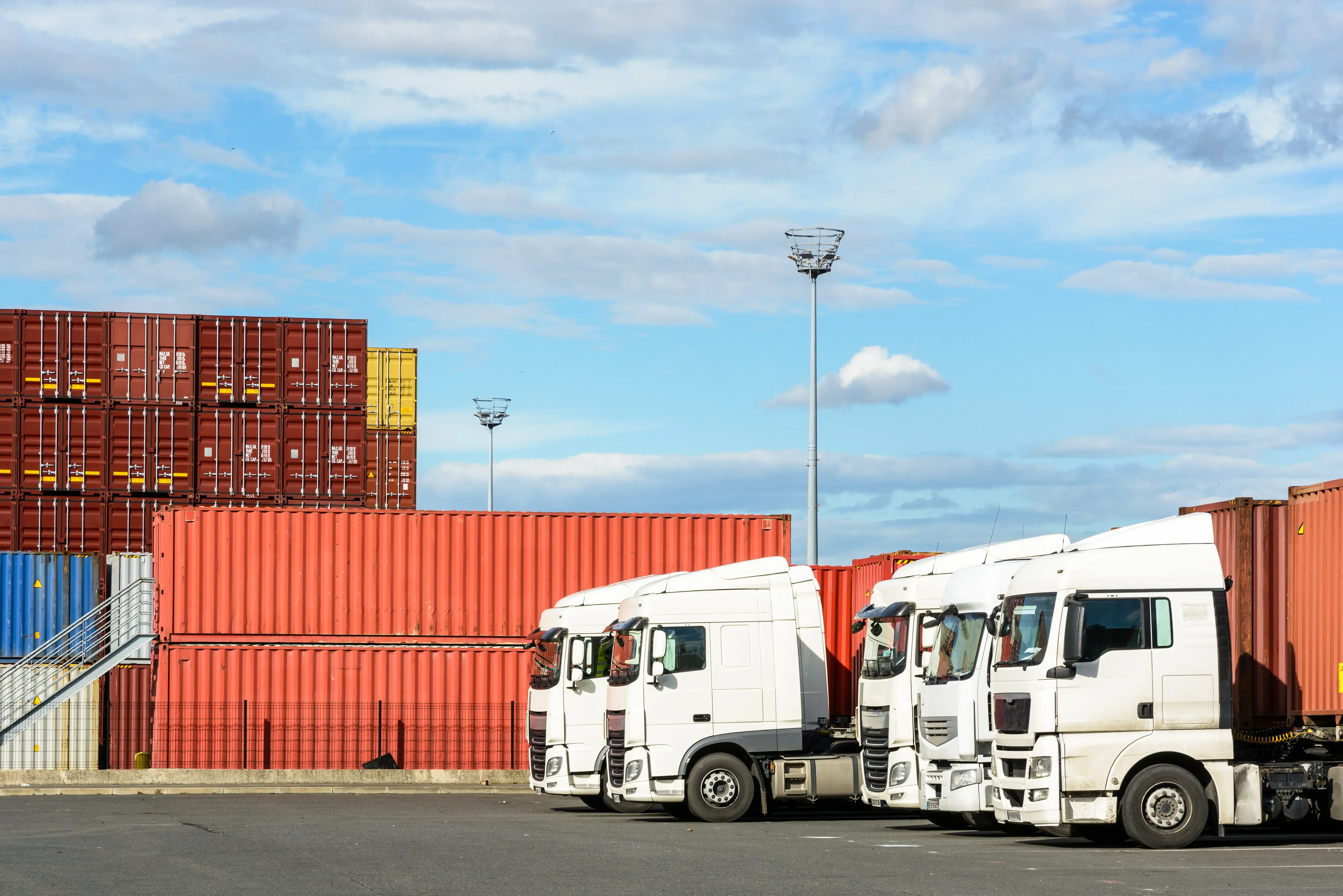 A row of white semi-trailer trucks parked on the container storage platform of the intermodal terminal of a river port with containers in the background