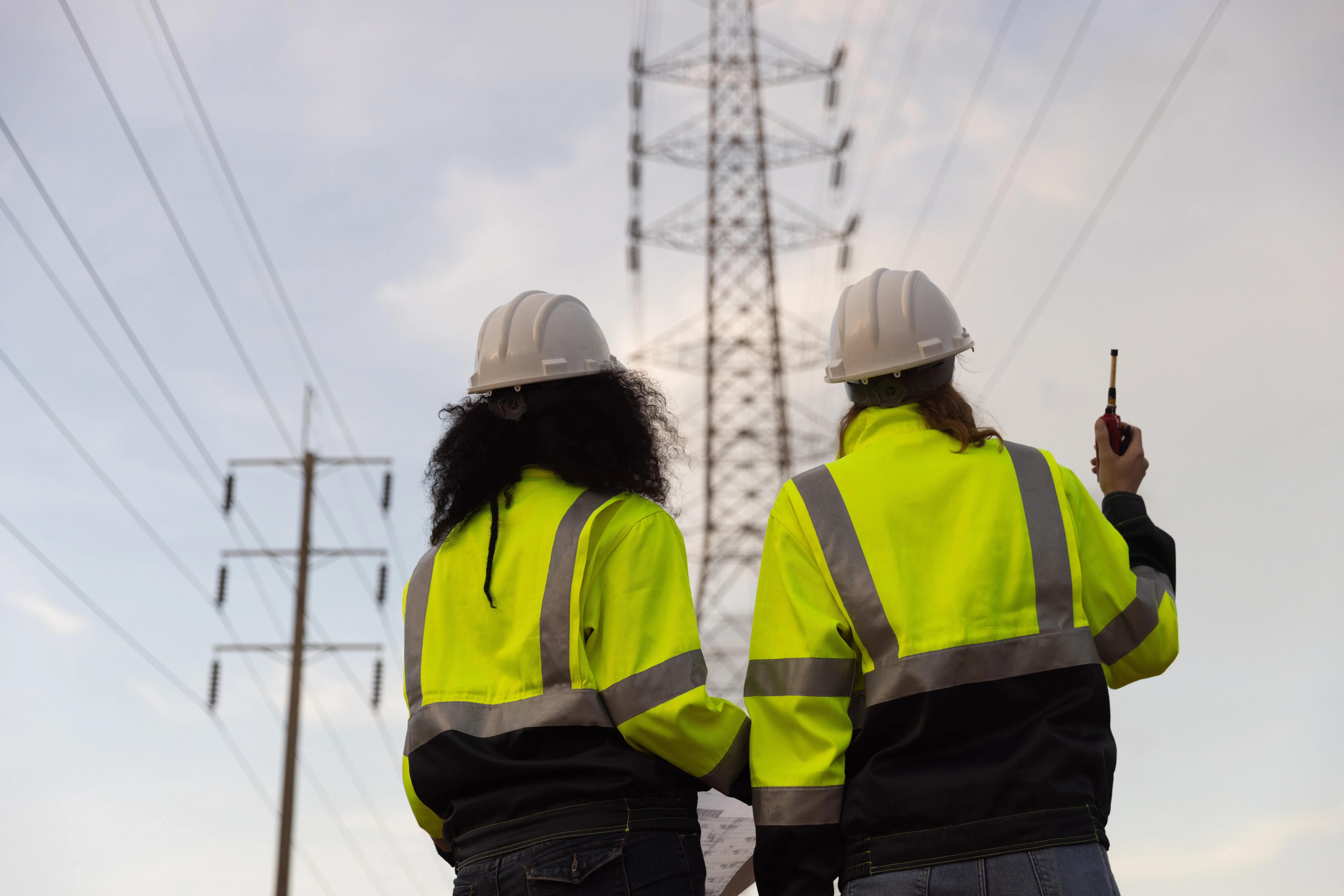 Back view of two female industry engineers working and discussing in front of a voltage-generating station and high-voltage pole