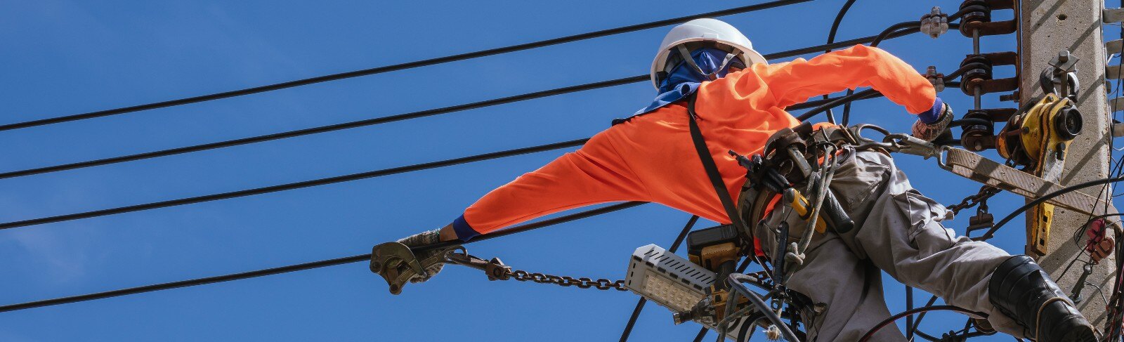 Electrician with safety equipment and work tools is installing cable lines and electrical system on electric power pole against blue sky