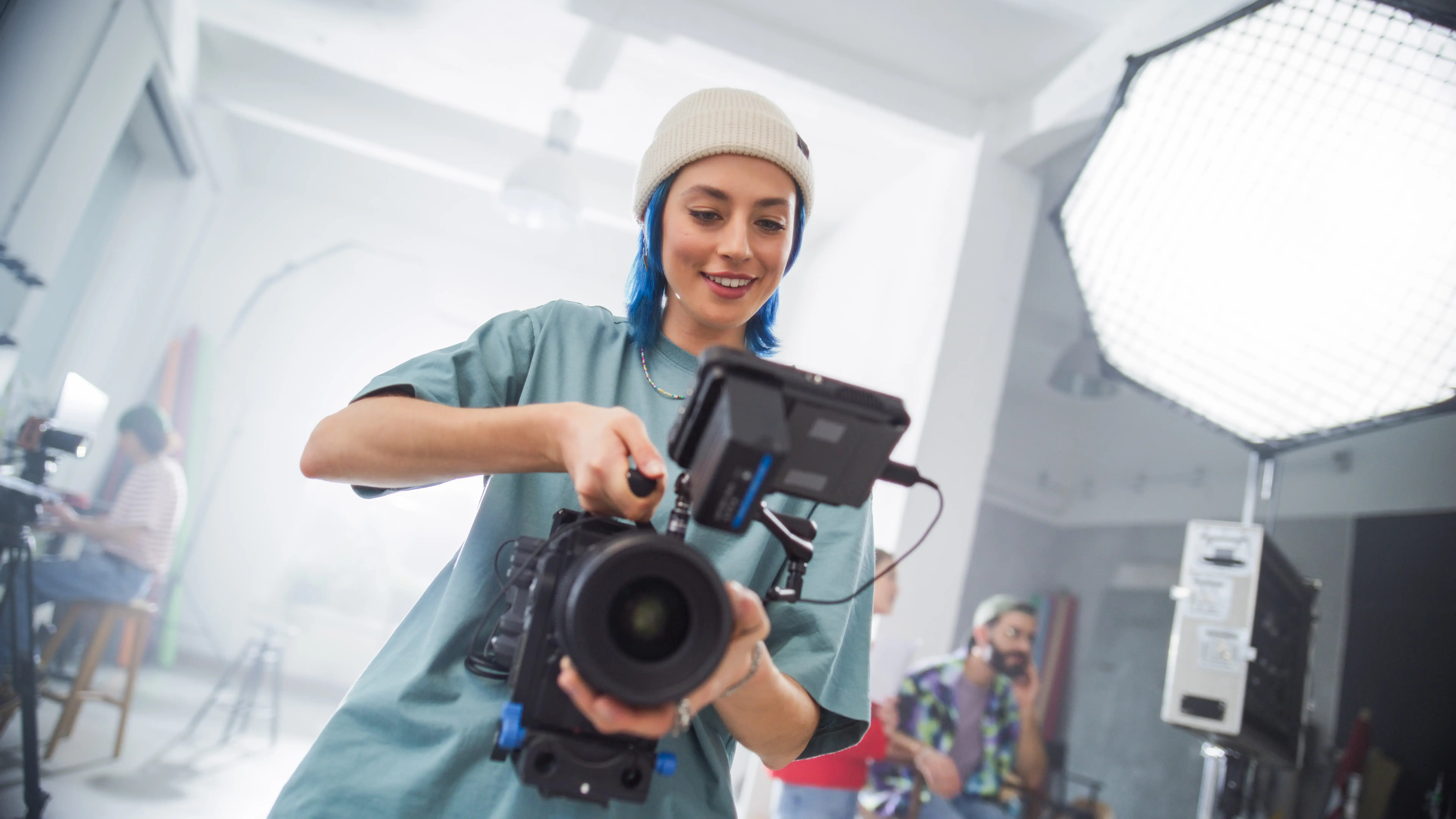 Young Caucasian Female Filmmaker In A Beanie Adjusts A Camera On A Busy Film Set, Capturing The Essence Of Modern Cinematography With Her Team In The Background, Focused On A Creative Project.